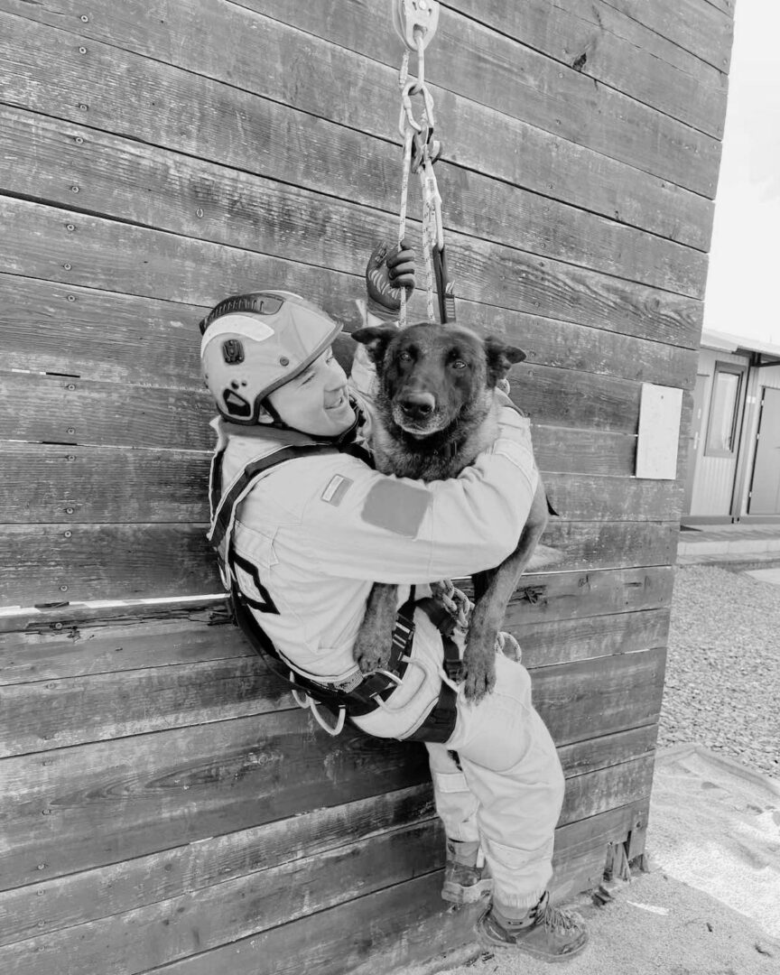 Climber in helmet and harness hugs a dog suspended by rope against a wooden wall surface.