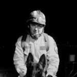 Firefighter kneeling beside a black German shepherd at night, both facing the camera, in turnout gear and helmet.
