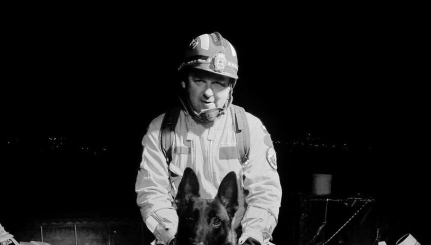Firefighter kneeling beside a black German shepherd at night, both facing the camera, in turnout gear and helmet.