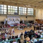 Indoor gymnasium hosting a majorettes competition; groups of young dancers in colorful outfits stand in rows on the court while audience sits in bleachers overseeing the event. Banner reads 'Puchar Polski Mażoretek'.