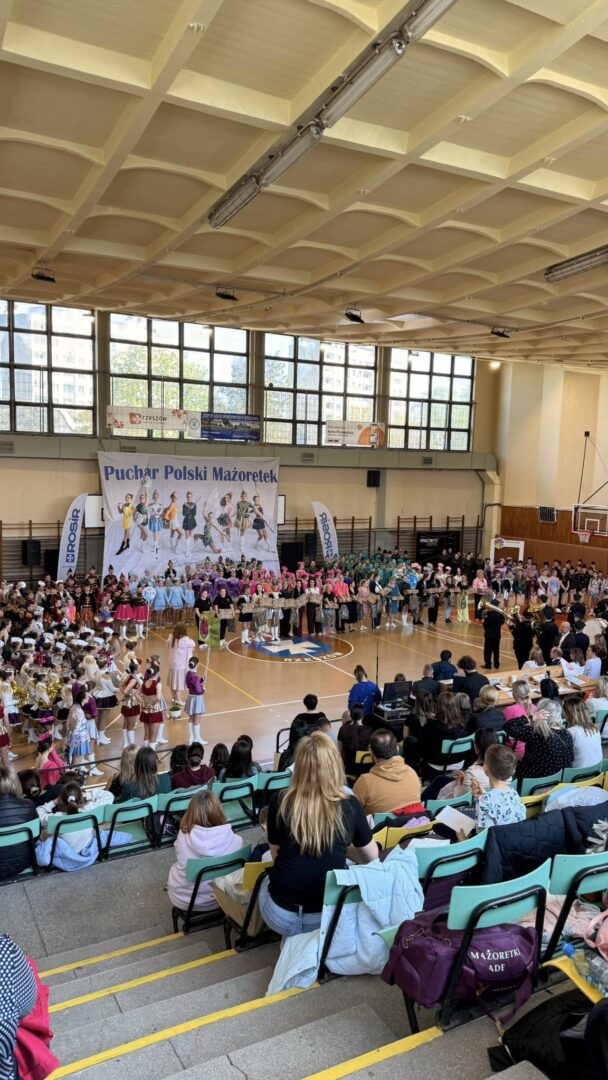 Indoor gymnasium hosting a majorettes competition; groups of young dancers in colorful outfits stand in rows on the court while audience sits in bleachers overseeing the event. Banner reads 'Puchar Polski Mażoretek'.