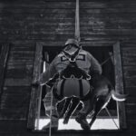 Firefighter in turnout gear climbs through a broken window of a weathered wooden building, rope trailing beside him, with a dog following nearby.