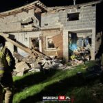 Firefighters in protective gear stand amid a collapsed brick house at night, with rubble and debris scattered outside.
