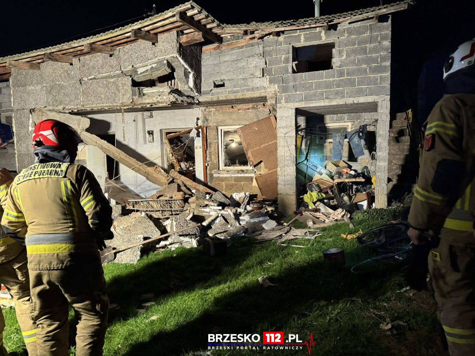 Firefighters in protective gear stand amid a collapsed brick house at night, with rubble and debris scattered outside.