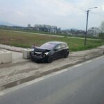 Black hatchback with front-end damage beside concrete barriers on a roadside; a man inspects the hood.