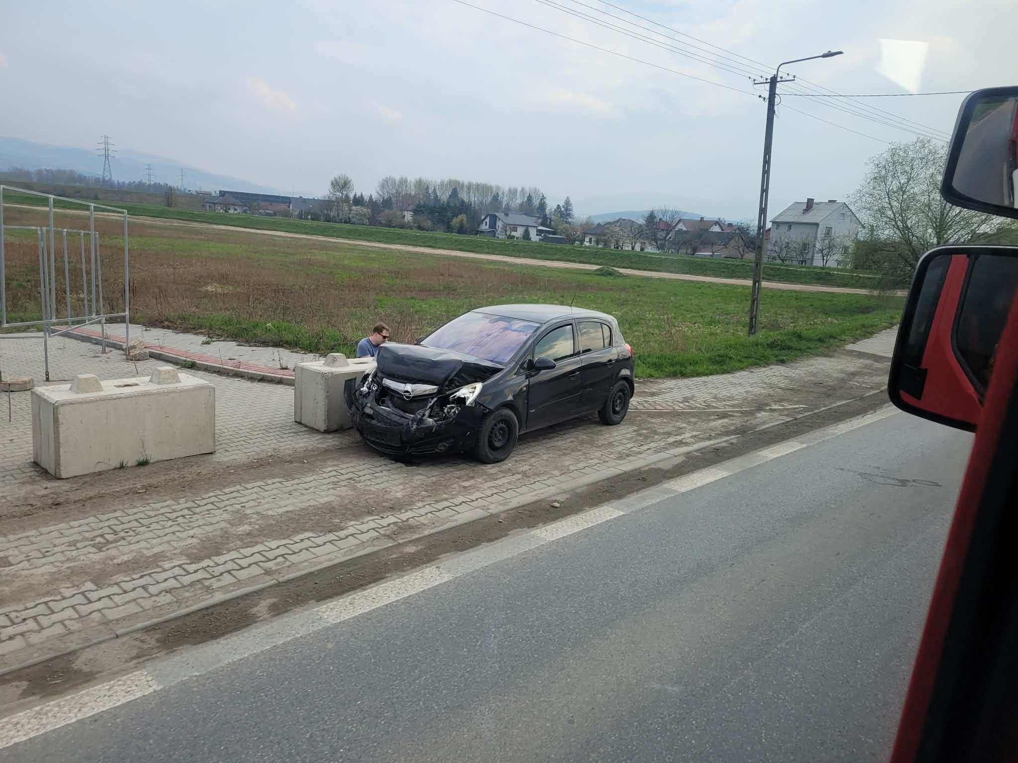 Black hatchback with front-end damage beside concrete barriers on a roadside; a man inspects the hood.