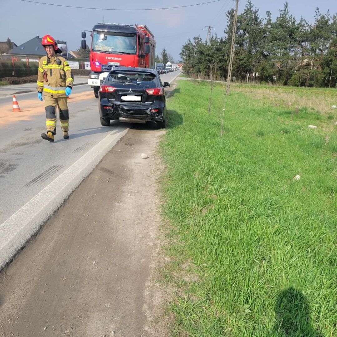 Firefighter in turnout gear walking past a damaged black car after a collision; a red fire truck is parked behind.