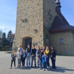 Group of children and two adults posing outside a tall stone church on a sunny day, with a cross structure nearby.
