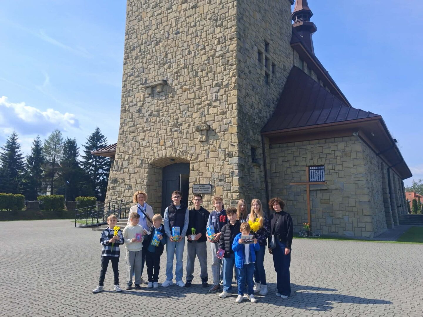 Group of children and two adults posing outside a tall stone church on a sunny day, with a cross structure nearby.