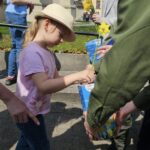 Young girl in a straw hat and pink shirt hands a flower to an adult during an outdoor event, with a basket of flowers nearby.