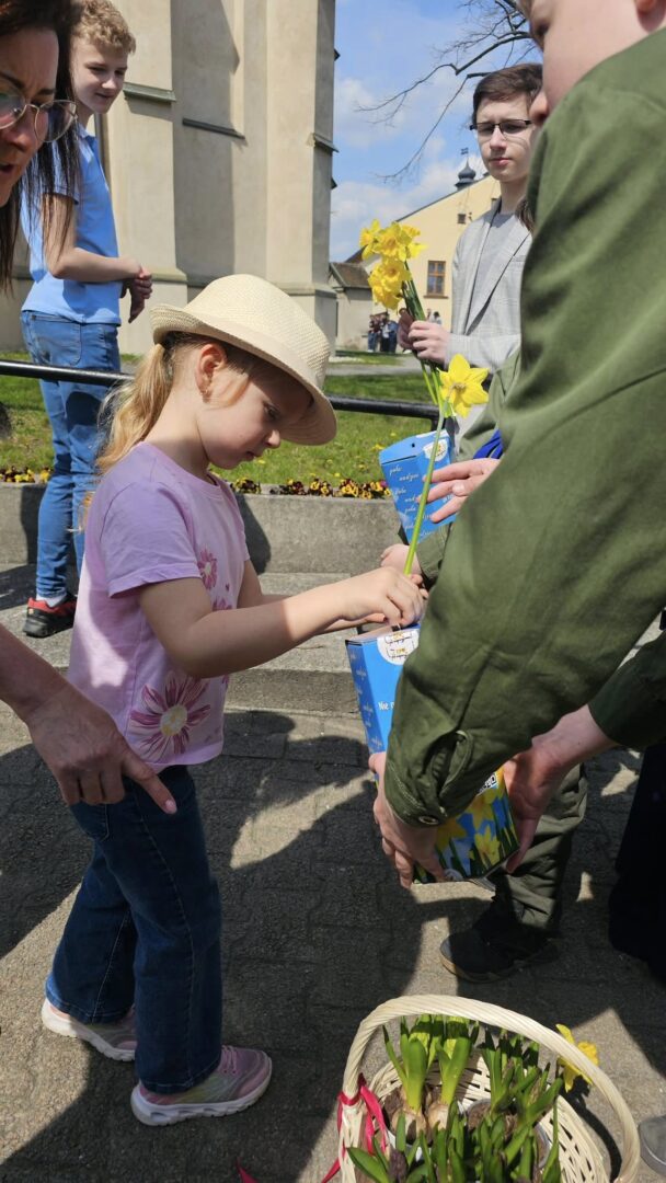 Young girl in a straw hat and pink shirt hands a flower to an adult during an outdoor event, with a basket of flowers nearby.
