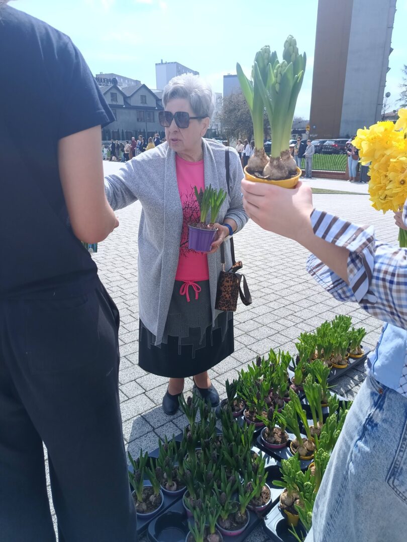 Older woman in pink shirt and gray cardigan, wearing sunglasses, holds a small purple pot with green shoots at an outdoor plant market. People and potted bulbs are around her.
