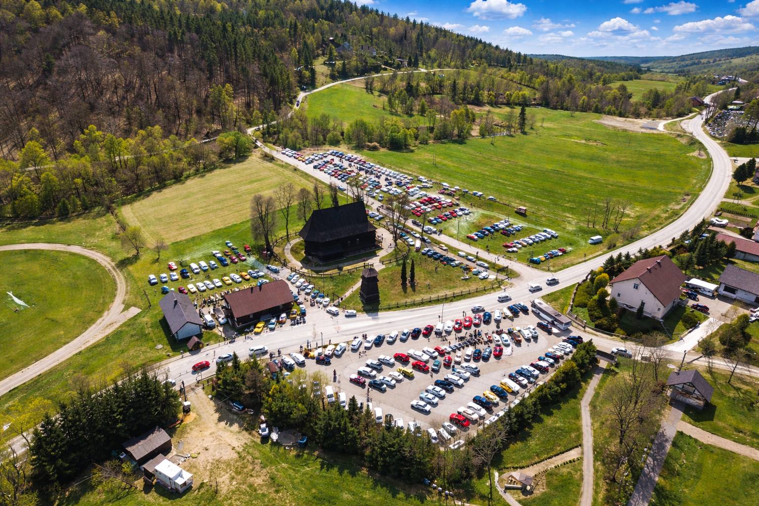 Aerial view of a rural church complex with a large parking lot full of colorful cars and green hills surrounding it on a sunny day.
