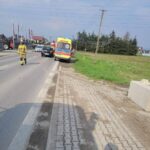 Emergency response scene on a two-lane road: a yellow ambulance on the shoulder, a firefighter in uniform walking, and a dark car nearby.