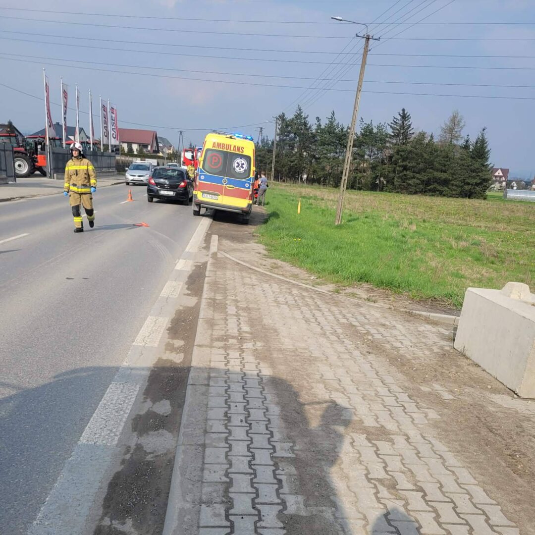 Emergency response scene on a two-lane road: a yellow ambulance on the shoulder, a firefighter in uniform walking, and a dark car nearby.