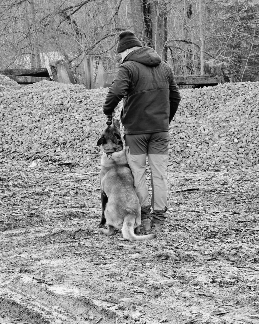 Man in a dark jacket and beanie stands beside a seated dog outdoors near a gravel pile and bare trees.