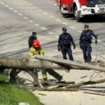 Firefighter in yellow gear and red helmet clears a large fallen tree beam while two police officers stand nearby on a sidewalk by a road with a crashed debris scene.