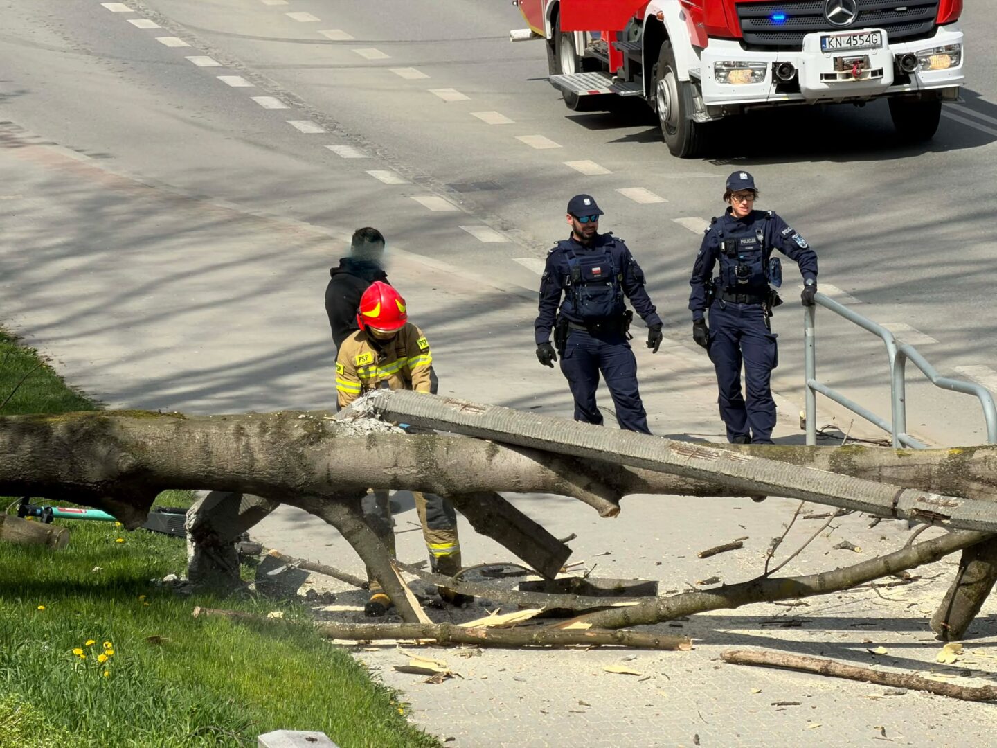 Firefighter in yellow gear and red helmet clears a large fallen tree beam while two police officers stand nearby on a sidewalk by a road with a crashed debris scene.