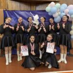 Eight dancers in matching black outfits pose with pink medals and diplomas during a celebration, pastel balloon arch to the right.