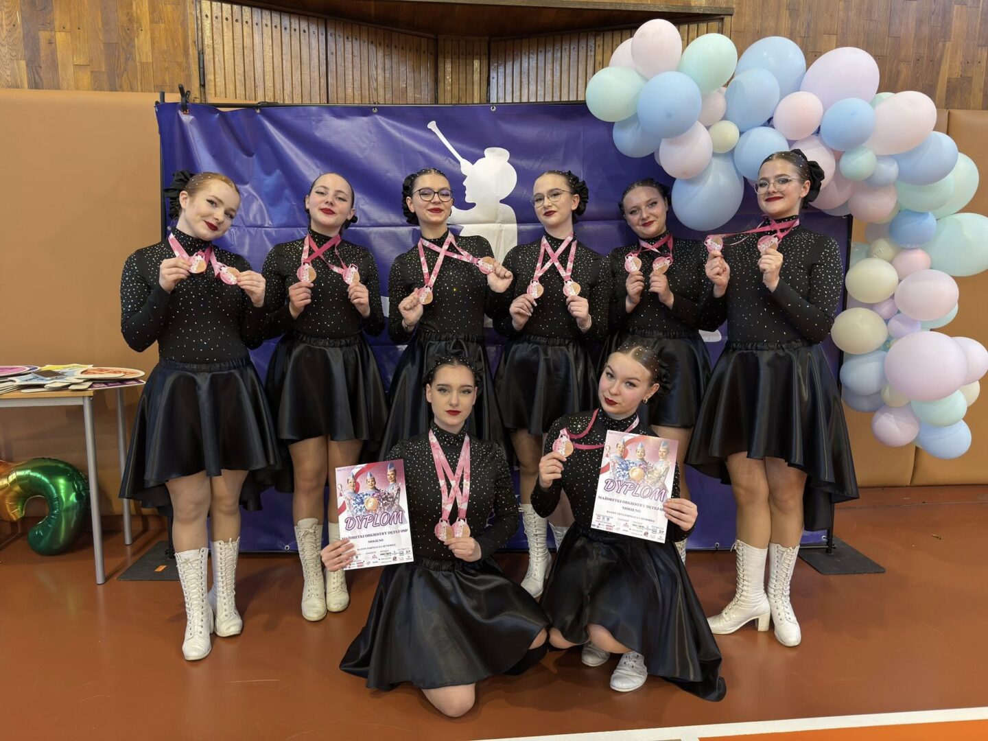 Eight dancers in matching black outfits pose with pink medals and diplomas during a celebration, pastel balloon arch to the right.