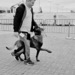 Man wearing a light shirt and jacket walks with a dog on a leash; the dog stands on its hind legs, looking up at him.