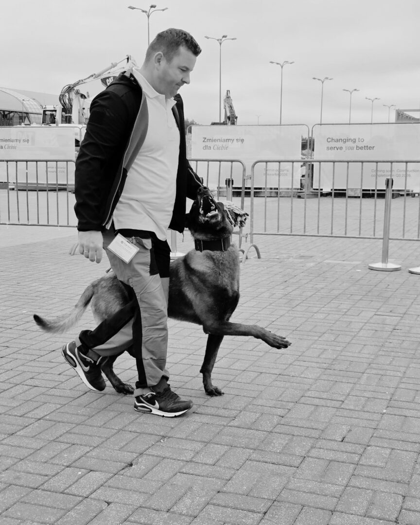 Man wearing a light shirt and jacket walks with a dog on a leash; the dog stands on its hind legs, looking up at him.