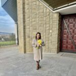 Smiling woman standing outside a beige stone building, holding a bouquet of yellow flowers in a beige coat and brown boots.