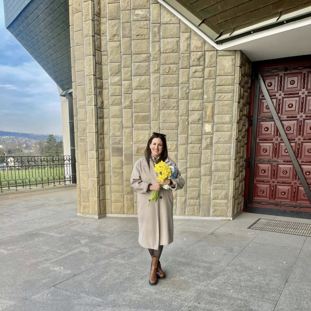 Smiling woman standing outside a beige stone building, holding a bouquet of yellow flowers in a beige coat and brown boots.