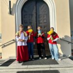 Four people stand on church steps in front of a large arched door; one person wears a white liturgical robe with a red robe and holds a wicker basket, while the others in red shirts hold yellow flowers and a card.