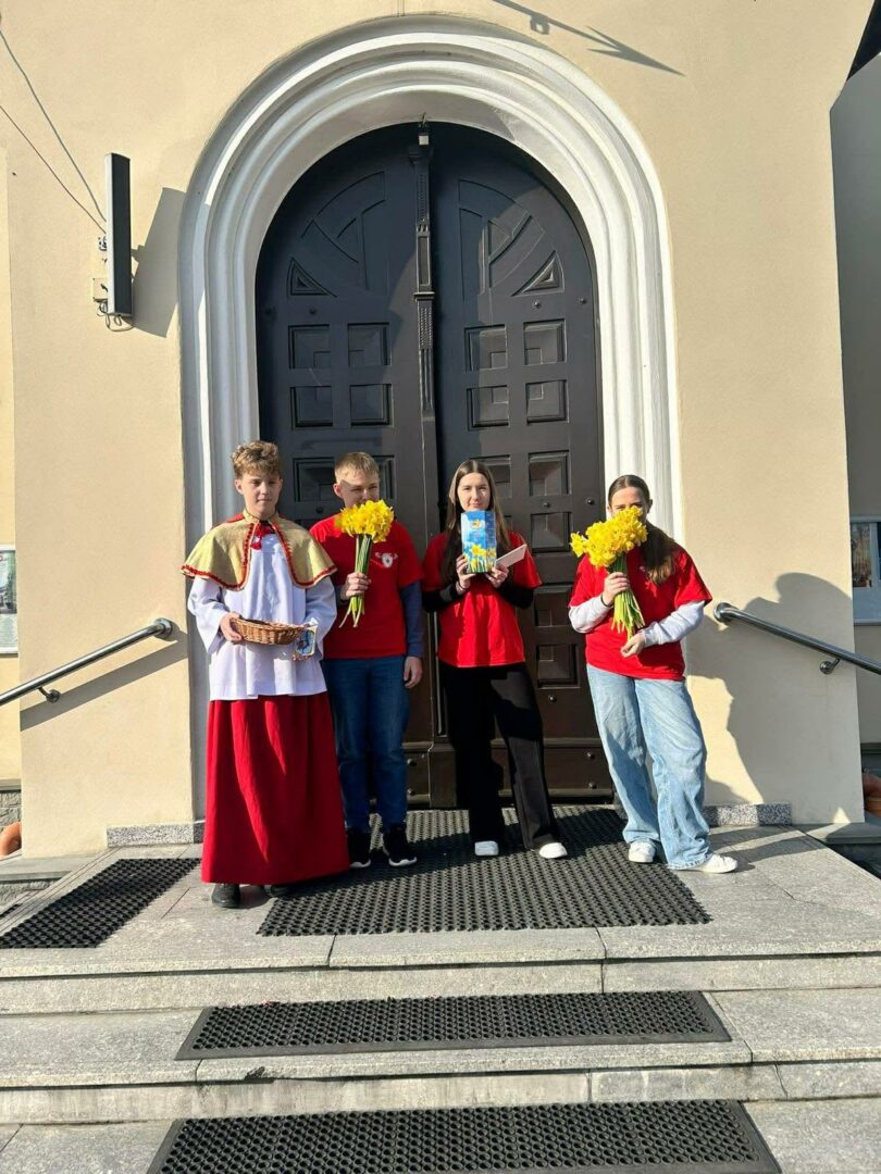 Four people stand on church steps in front of a large arched door; one person wears a white liturgical robe with a red robe and holds a wicker basket, while the others in red shirts hold yellow flowers and a card.