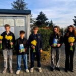 Five youths stand outdoors in a row, each holding a yellow daffodil bouquet; two hold blue gift boxes with bright designs. They pose in front of a bulletin board and greenery on a sunny day.