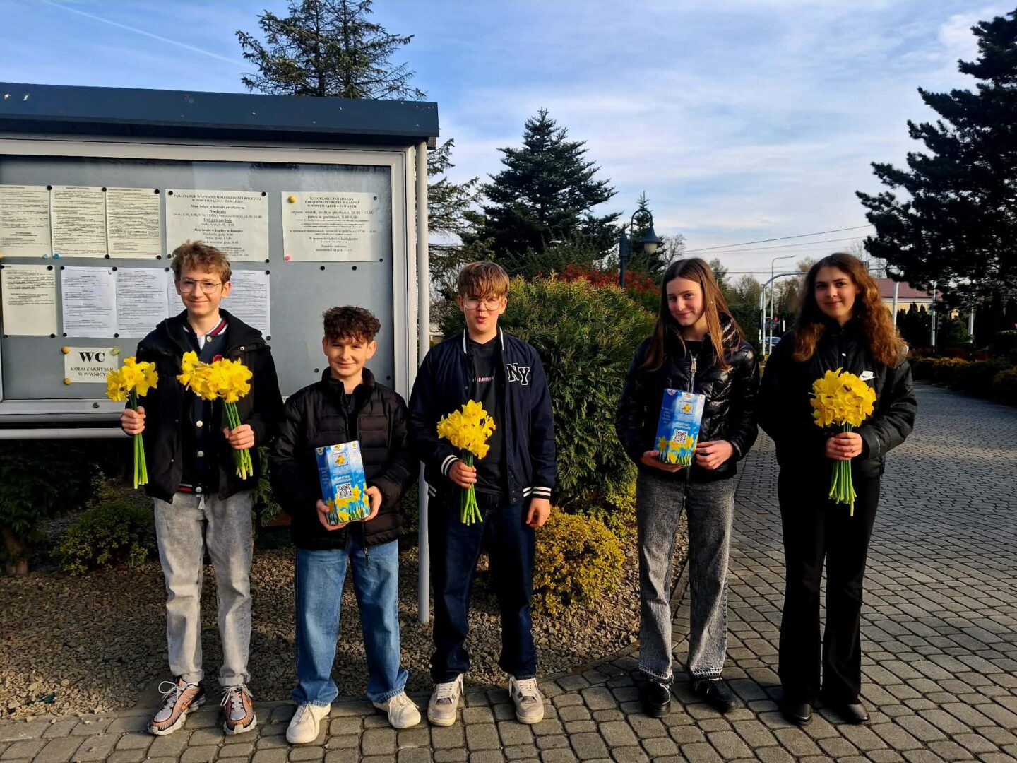Five youths stand outdoors in a row, each holding a yellow daffodil bouquet; two hold blue gift boxes with bright designs. They pose in front of a bulletin board and greenery on a sunny day.