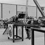 German Shepherd balancing on a raised obstacle during a public dog demonstration, with a handler nearby and spectators behind a barrier.
