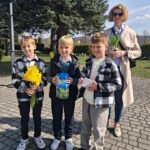 Woman and three boys outdoors on a sunny day; one boy holds yellow daffodils, another carries a blue gift/foam can, and the third holds a small card, with a supervising woman holding a potted plant in the background.