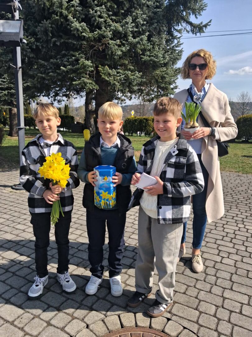 Woman and three boys outdoors on a sunny day; one boy holds yellow daffodils, another carries a blue gift/foam can, and the third holds a small card, with a supervising woman holding a potted plant in the background.