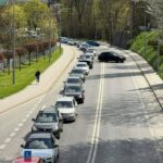 Line of cars in a queue on a curved road, led by a red learner car with an L plate; a pedestrian walks on the sidewalk beside a park.