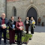 Five adults stand outside a stone church, each holding a card or small bouquet, with yellow tulips in neat pots along the path in front of the entrance.