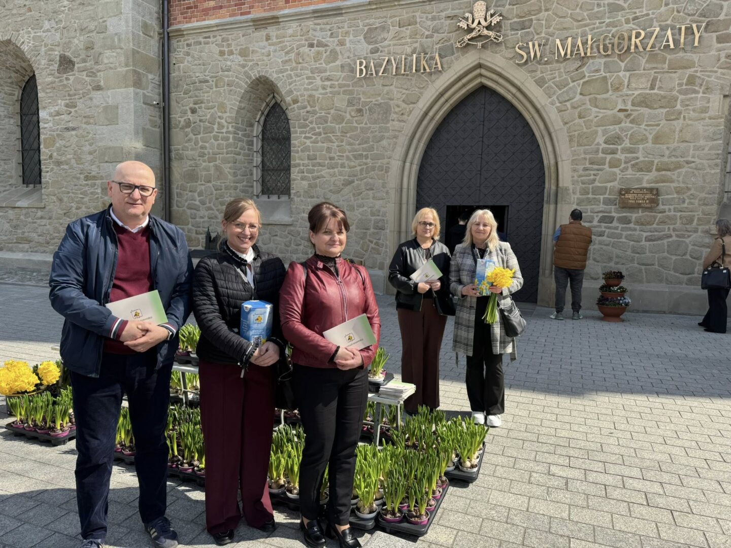 Five adults stand outside a stone church, each holding a card or small bouquet, with yellow tulips in neat pots along the path in front of the entrance.