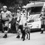 Rescue workers in protective suits walk with a police/hd dog on a leash near a white van in an outdoor scene.