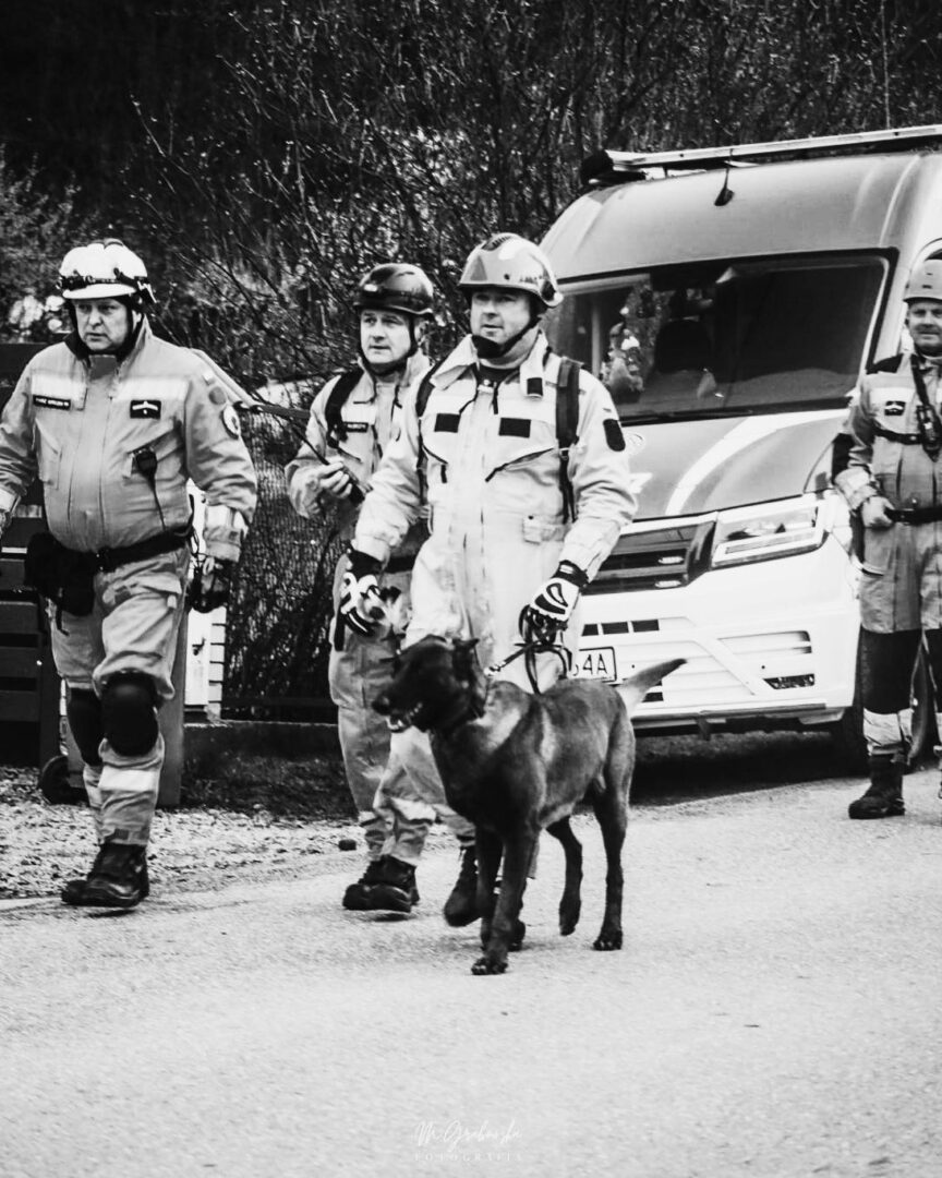 Rescue workers in protective suits walk with a police/hd dog on a leash near a white van in an outdoor scene.