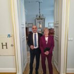 Two professionally dressed people stand together in a doorway of a formal, light-blue hallway with a chandelier overhead.