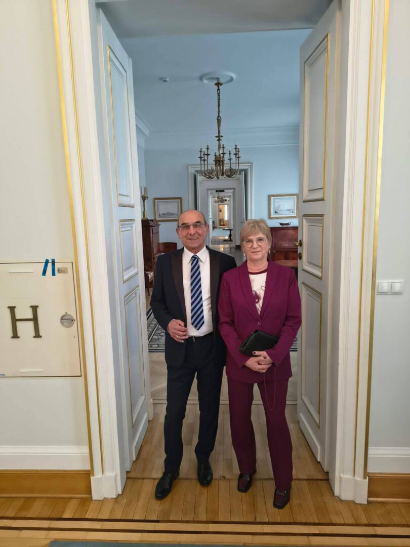 Two professionally dressed people stand together in a doorway of a formal, light-blue hallway with a chandelier overhead.