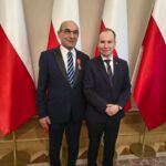 Two men in dark suits posing indoors with Polish flags in the background; one wears a medal on his lapel.