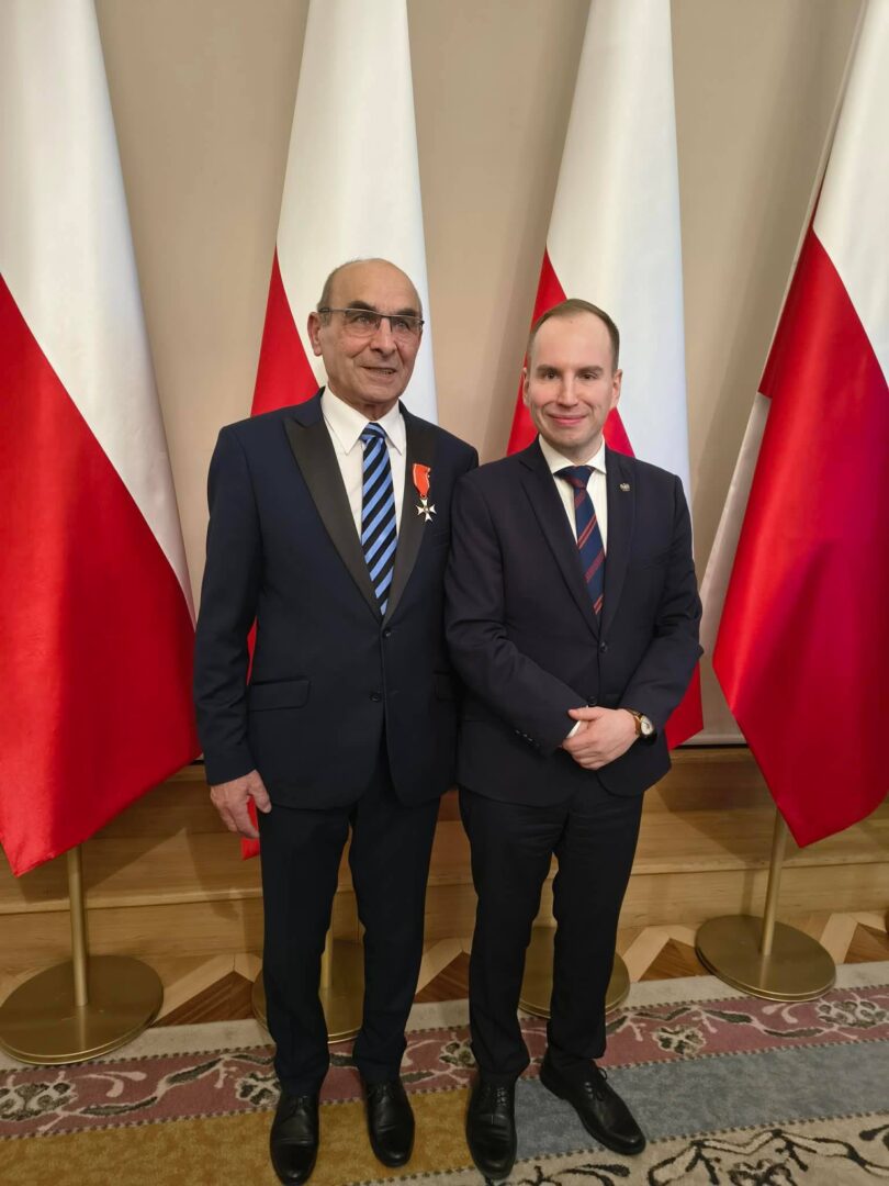 Two men in dark suits posing indoors with Polish flags in the background; one wears a medal on his lapel.