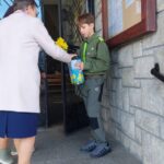 Woman in a light coat hands a yellow daffodil to a boy scout in a green uniform outside a stone building, who also holds a blue gift bag.