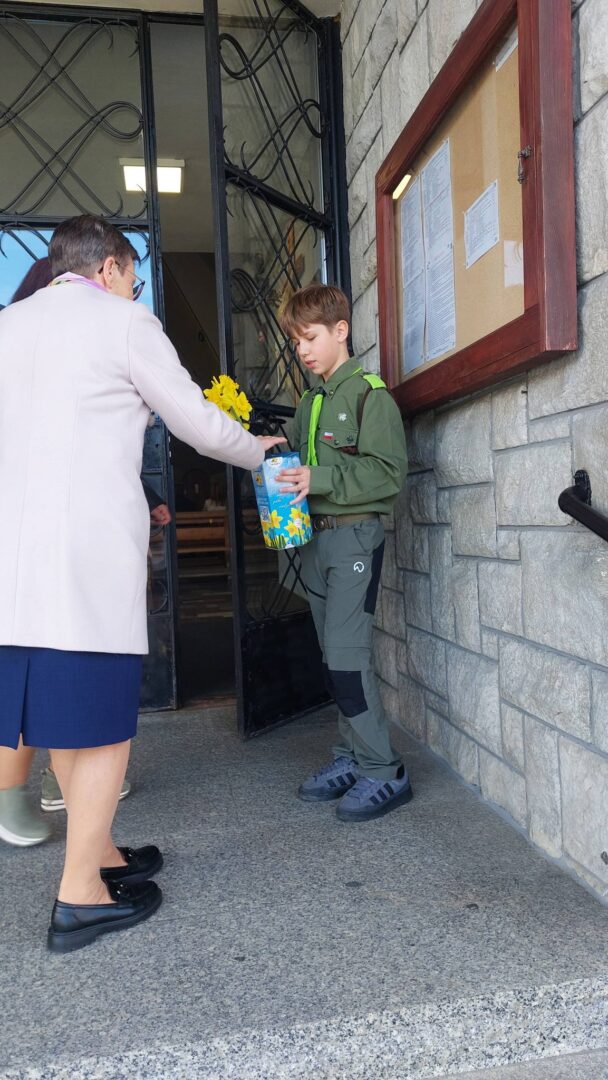 Woman in a light coat hands a yellow daffodil to a boy scout in a green uniform outside a stone building, who also holds a blue gift bag.