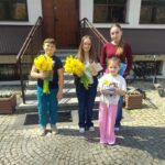 Four children pose outside a building, each holding bright yellow daffodils, with a doorway and stairs behind them.