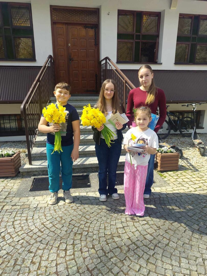 Four children pose outside a building, each holding bright yellow daffodils, with a doorway and stairs behind them.
