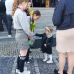 Young girl in white tights hands a bouquet of yellow tulips to two teenage girls in gray uniforms outside a building, while others wait on steps.