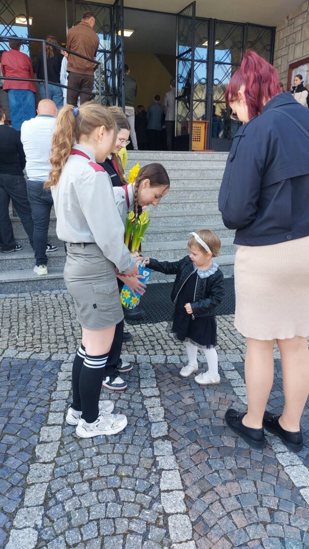 Young girl in white tights hands a bouquet of yellow tulips to two teenage girls in gray uniforms outside a building, while others wait on steps.